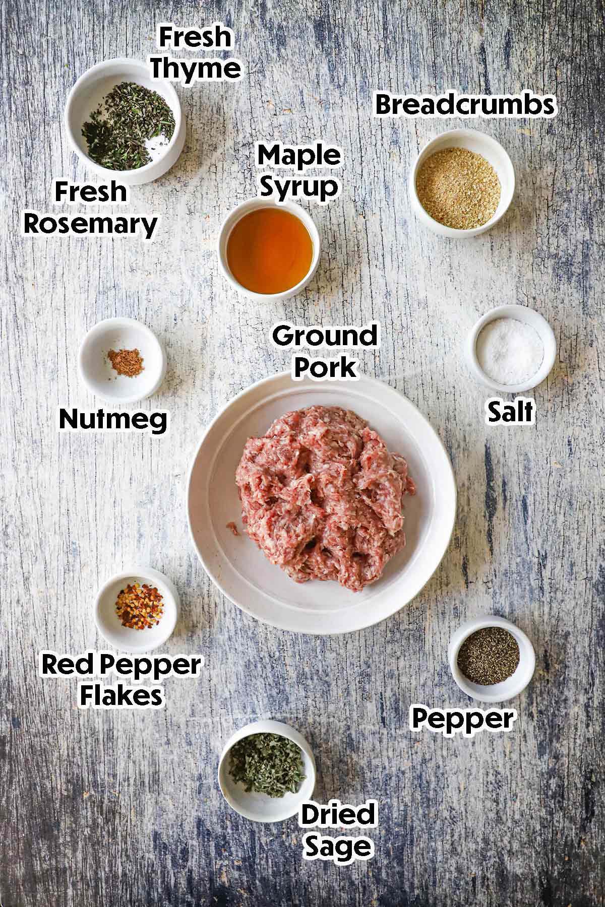 An arrangement of ingredients for maple sausage patties on a grey wooden background including ground pork, maple syrup, breadcrumbs, red pepper flakes, herbs, and seasonings.