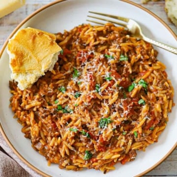 An overhead view of a shallow pasta bowl filled with a serving of One-Pot Ground Beef Orzo with a torn piece of Italian bread resting on the side of the bowl.