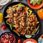 An overhead view of a large fajita skillet filled with a large serving of best shrimp fajitas along with sautéed strips of colorful bell peppers all surrounded by bowls of pico de gallo, cotija cheese, and lime wedges.