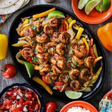 An overhead view of a large fajita skillet filled with a large serving of best shrimp fajitas along with sautéed strips of colorful bell peppers all surrounded by bowls of pico de gallo, cotija cheese, and lime wedges.