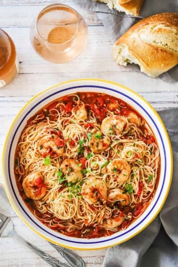 An overhead view of a large shallow pasta bowl filled with shrimp marinara and is garnished with chopped Italian parsley with a torn piece of Italian bread and two glasses of wine nearby.