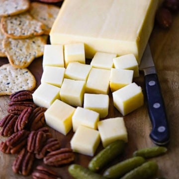 A close-up view of smoked mozzarella that has partially been cut into cubes and is resting on a board next to crackers, pecans, and baby dill pickles.