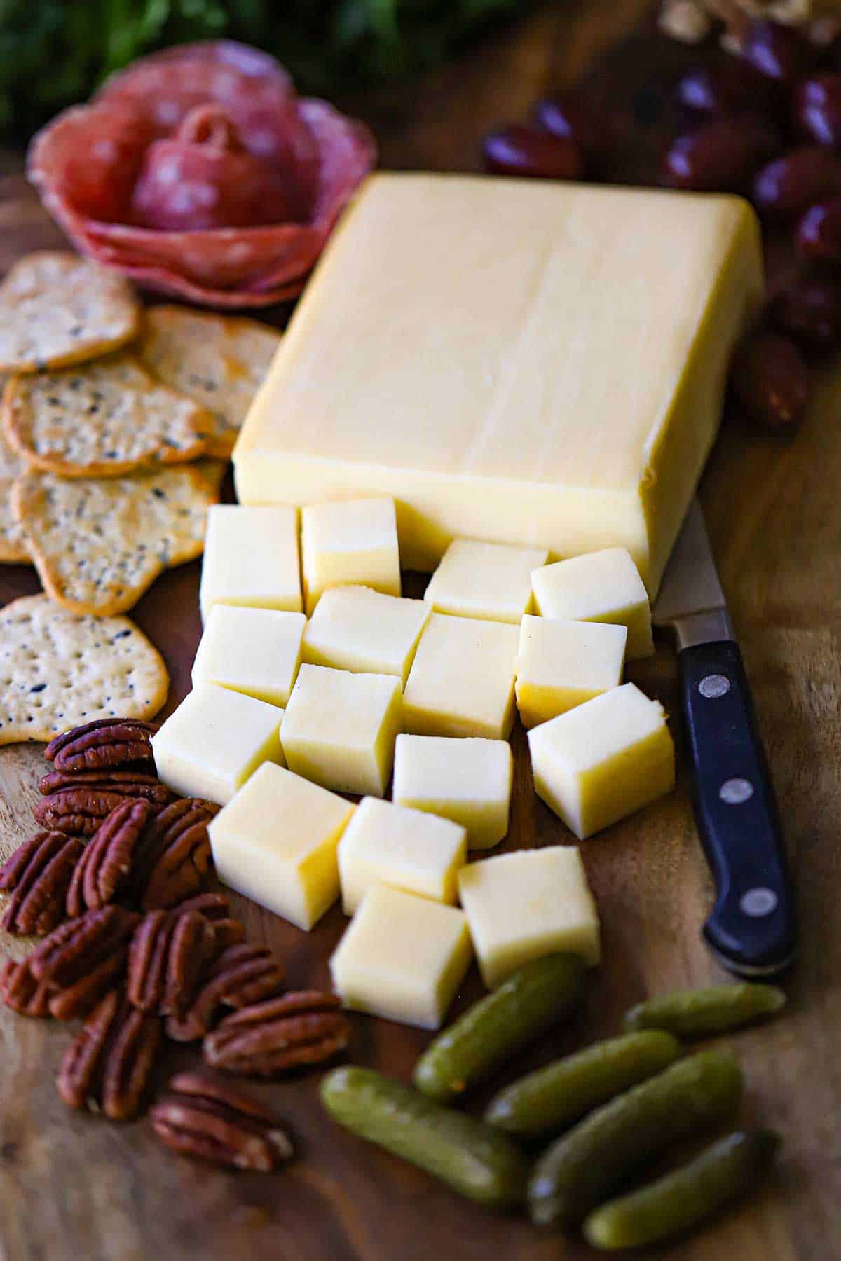 A straight-on view of smoked mozzarella that has partially been cut into cubes and is resting on a board next to crackers, pecans, salami, and baby dill pickles.