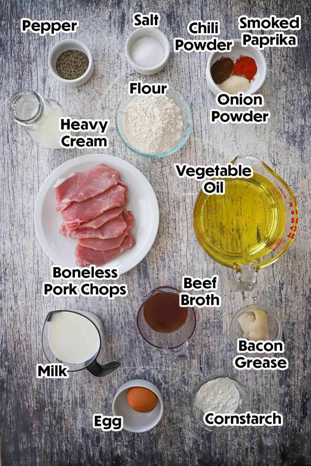 An arrangement of ingredients for Southern-Fried Pork Chops with Cream Gravy on a grey wooden background including boneless pork chops, flour, cream, oil, milk, beef broth, bacon grease, an egg, cornstarch, and seasonings.