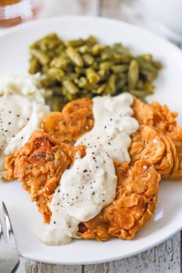 A straight-on view of two Southern-Fried Pork Chops with Cream Gravy on a white dinner plate along with a serving of green beans and mashed potatoes.