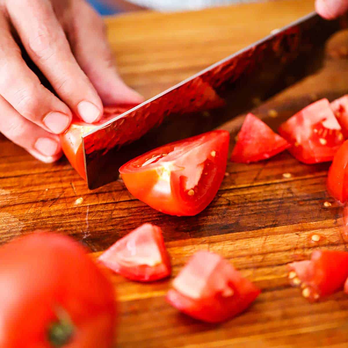 A person using a large knife to roughly chop a whole tomato on a wooden cutting board.