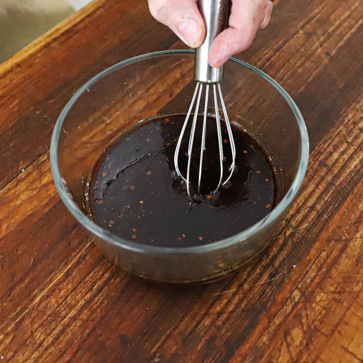 A person using a small whisk to combine the ingredients in a glass bowl for a Chinese kung pao sauce on wooden cutting board.