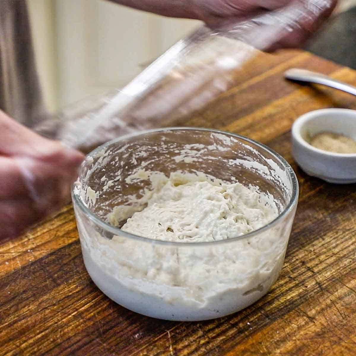 A person removing plastic wrap from the top of a glass bowl that is filled with poolish on a wooden cutting board.