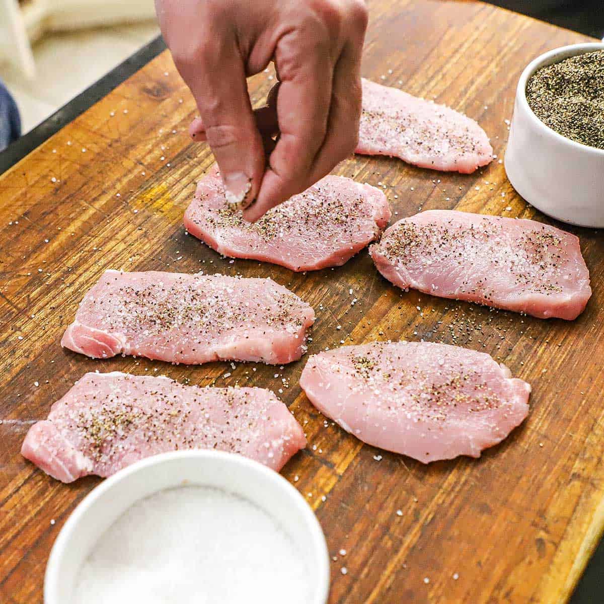 A person using his fingers to sprinkle salt and pepper over the tops of thin boneless pork chops on a wooden cutting board.