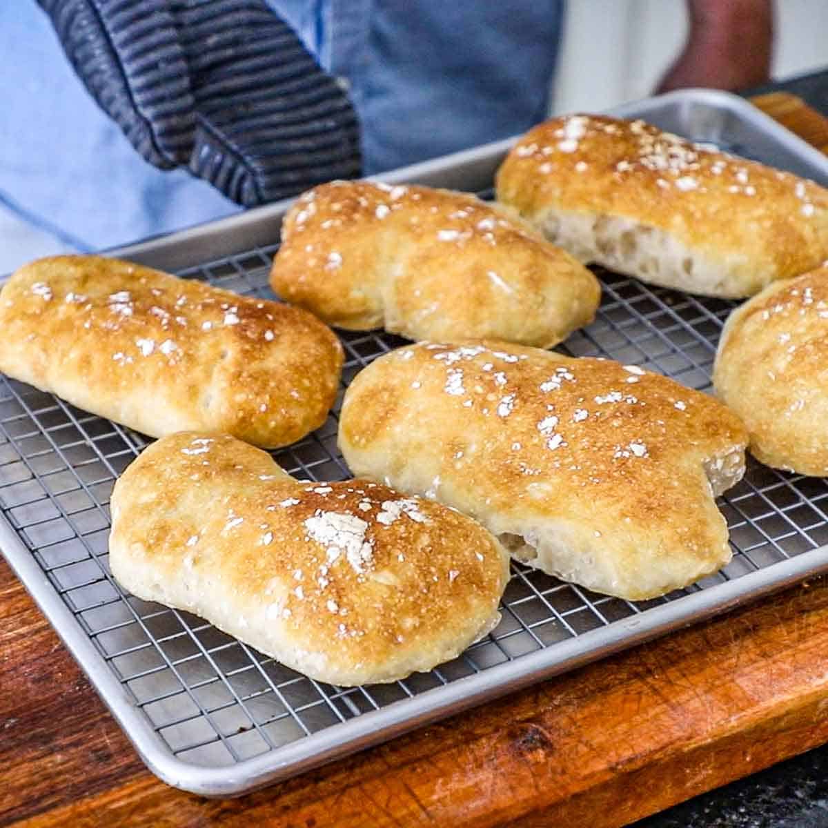 A large baking sheet lined with a baking rack containing six freshly baked homemade ciabatta rolls on a wooden cutting board.