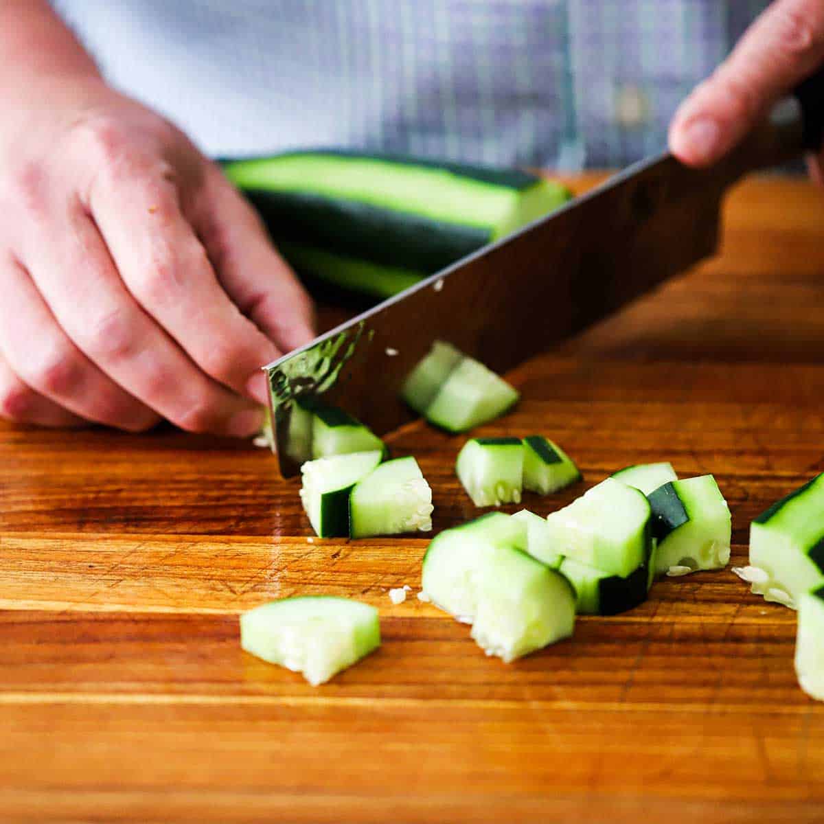 A person using a large knife to cut a whole cucumber into bite-sized pieces on a wooden cutting board.