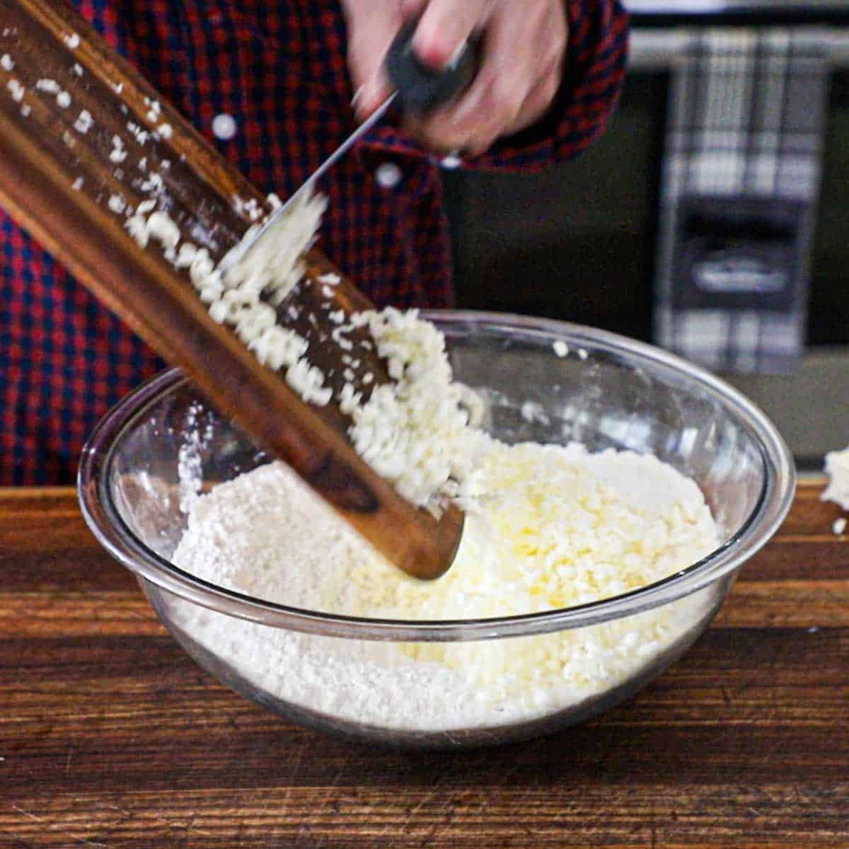A person using a pastry bench scraper to transfer chilled grated butter from a small cutting board into a large glass bowl filled with flour, baking powder, and baking soda.