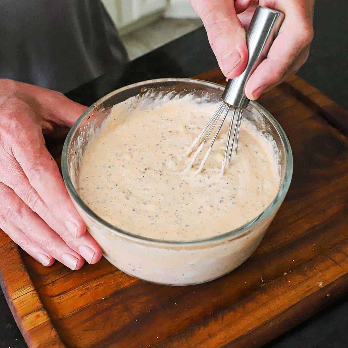 A person using a small whisk to stir a Cajun mayonnaise-based dressing in a small glass bowl on a wooden cutting board.