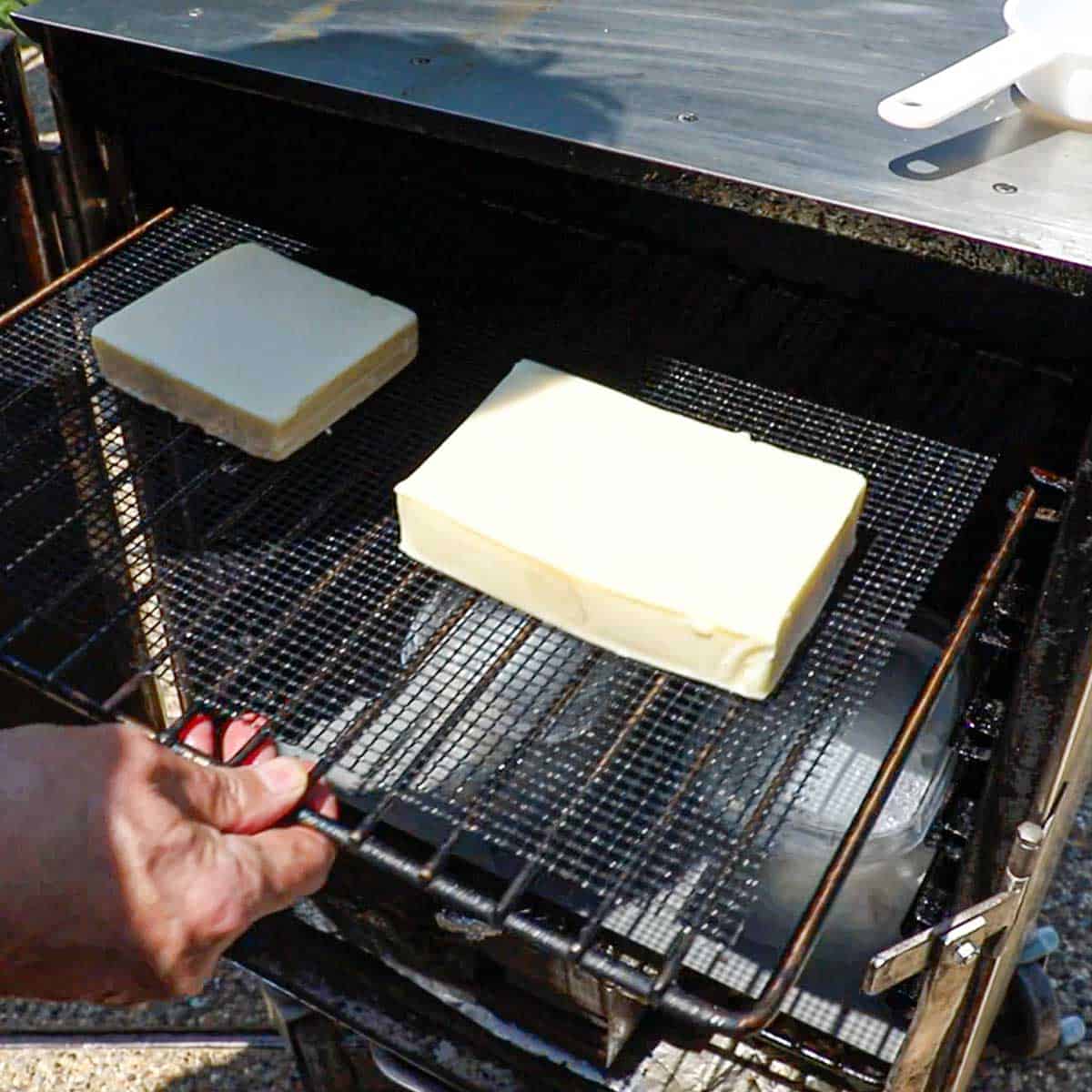 A person holding a non-stick mesh mat holding two blocks of cheese on the top level on the inside of an electric smoker.