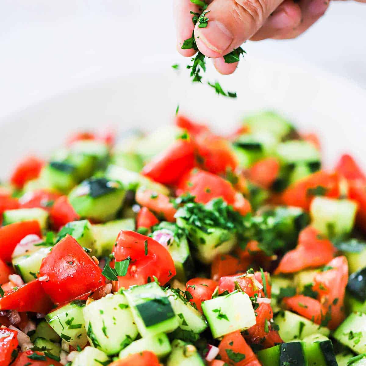 A person sprinkling chopped fresh Italian parsley over the top of a chopped Cucumber Tomato Salad in a large oval platter.