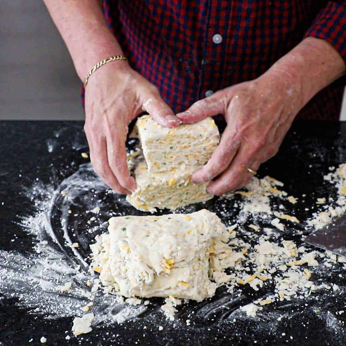 A person using his hands to stack to squares of uncooked flaky cheddar biscuit dough on a floured marble counter.