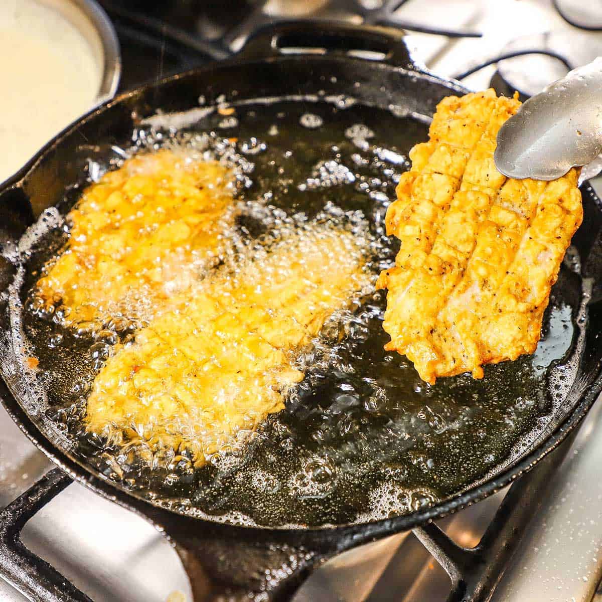 A person using a pair of metal tongs to lift a Southern-fried pork chop from a cast-iron skillet filled with hot oil and two other fried pork chops.