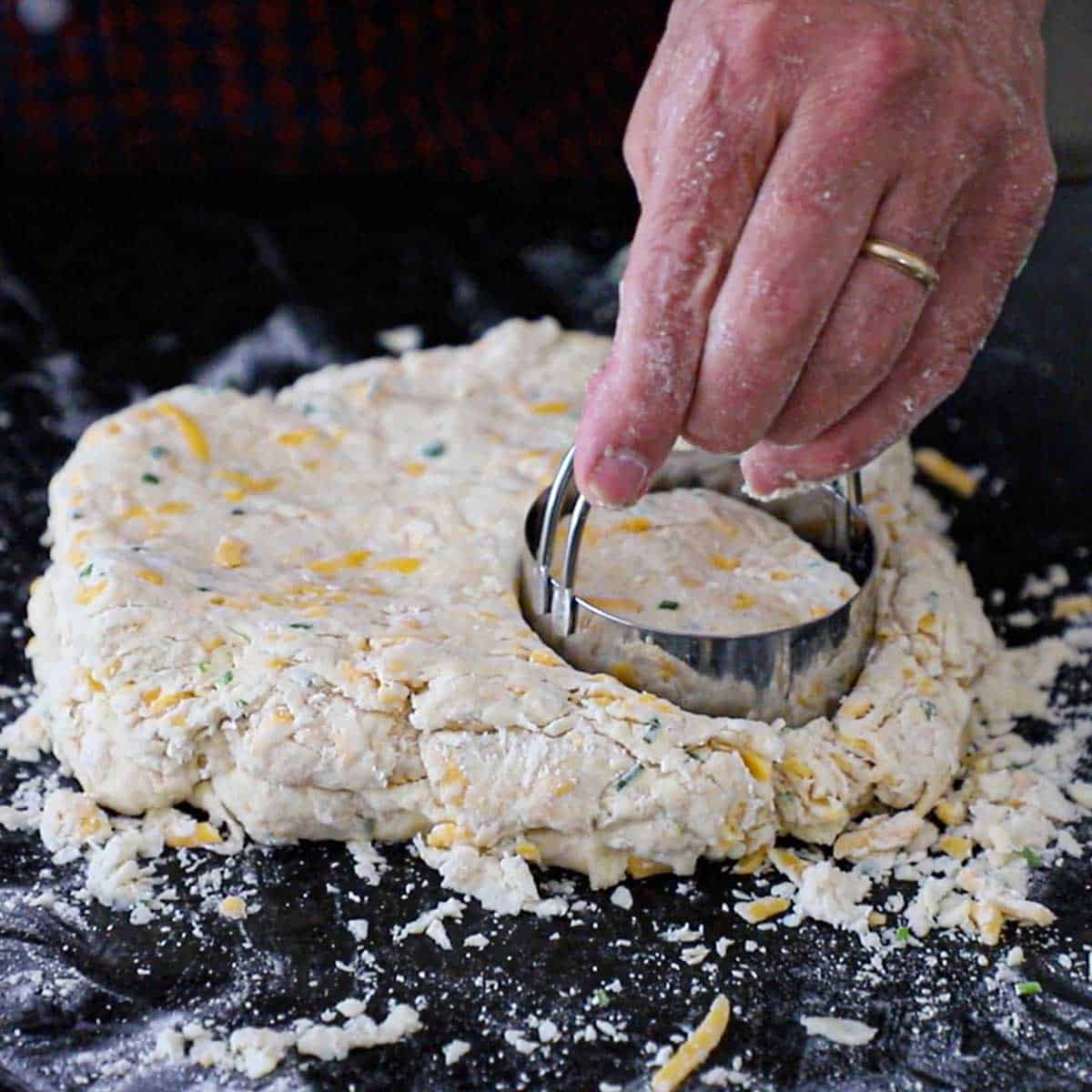A person pressing a round biscuit cutter into a square piece of cheddar biscuit dough on a flour marble counter.