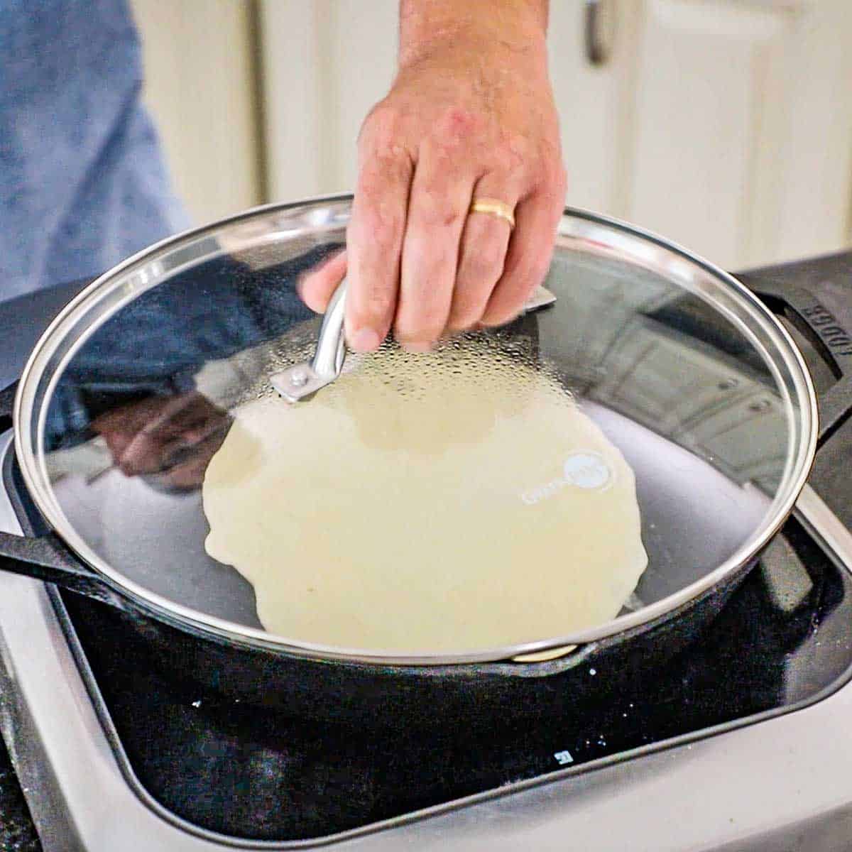 A person placing a clear lid over the top of a large cast-iron skillet with homemade naan in the hot skillet on an induction oven.