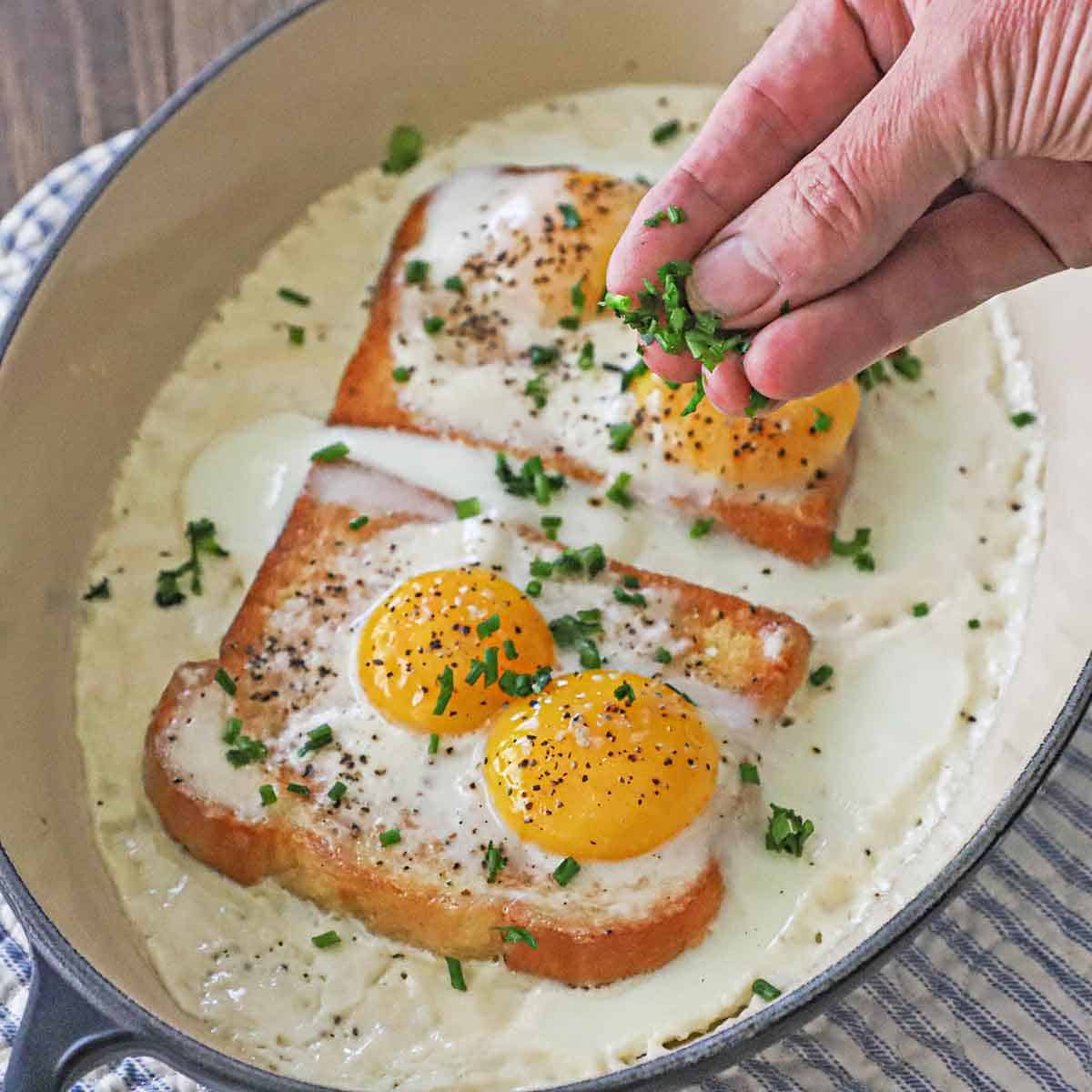 A person using his fingers to sprinkle chopped chives over two French Baked Eggs with Cream in an oval baking dish.