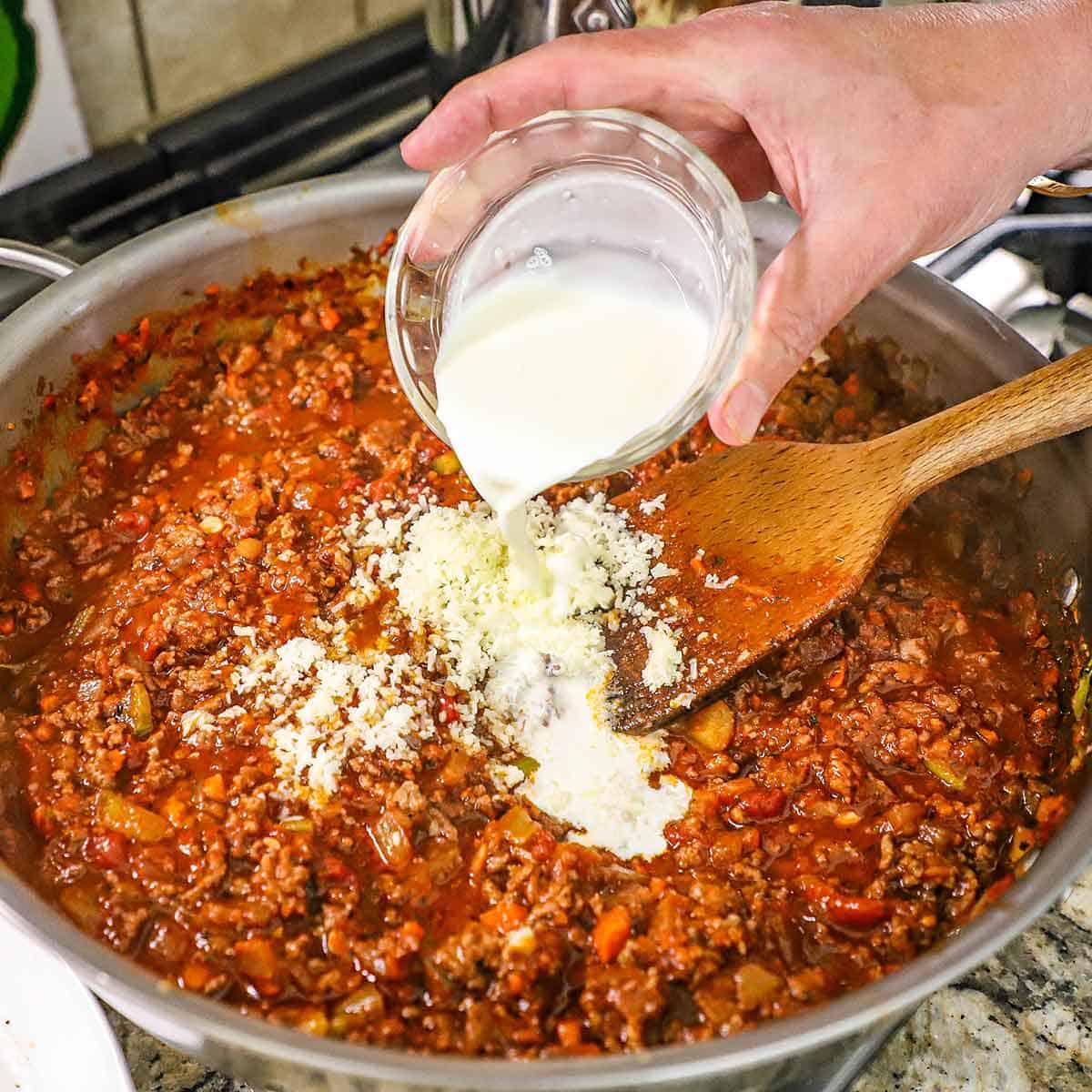 A person pouring heavy cream from a small bowl into a large skillet filled with a simmering bolognese sauce on a gas stove.