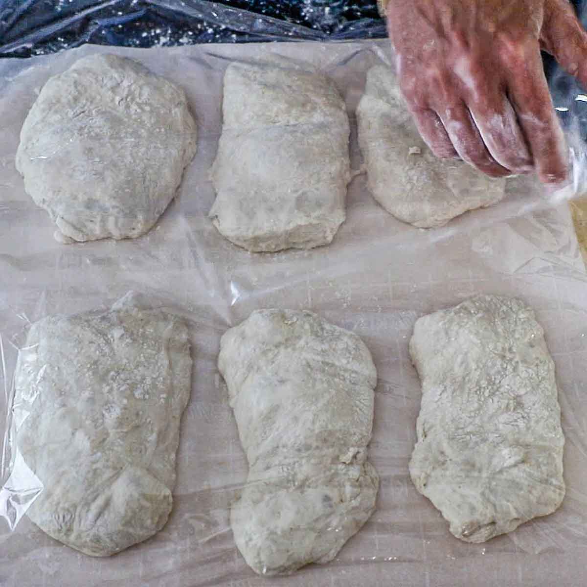 A person placing plastic wrap over the top of six uncooked mounds of ciabatta roll dough resting on parchment paper.