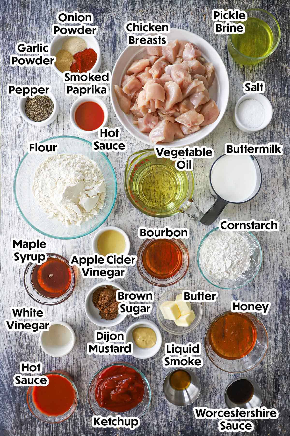 An arrangement of ingredients for crispy boneless chicken wings on a grey wooden background including chicken breast pieces, flour, cornstarch, buttermilk, pickle brine, butter, honey, maple syrup, Bourbon, hot sauce, and seasonings.