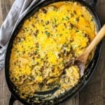 An overhead view of a large oval casserole dish filled with a cheesy potato hamburger casserole with a portion of it missing and a wooden spoon resting in the pan.