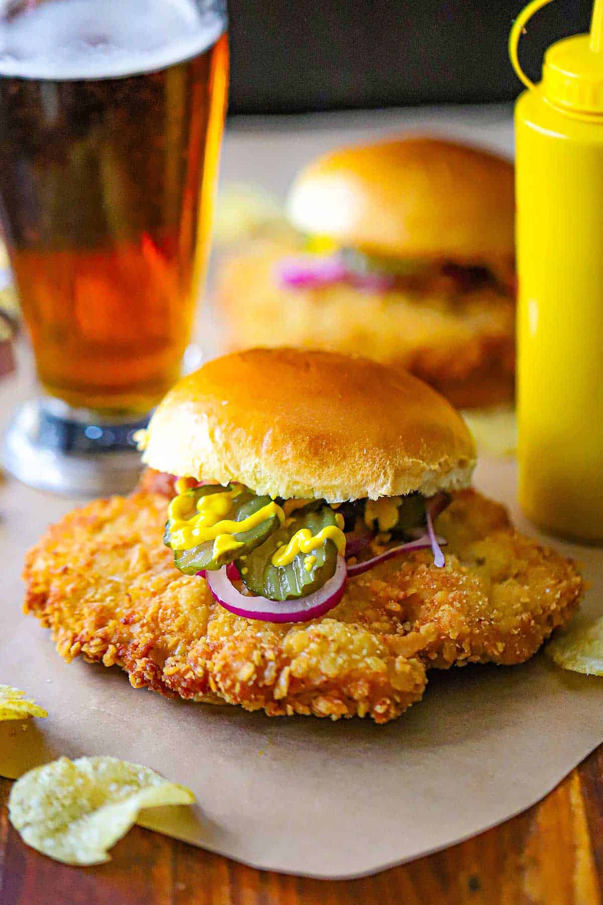 A straight-on view of a breaded pork tenderloin sandwich including a crispy pork tenderloin red onion, pickles, yellow mustard, all on a hamburger bun with a bottle of yellow mustard nearby.