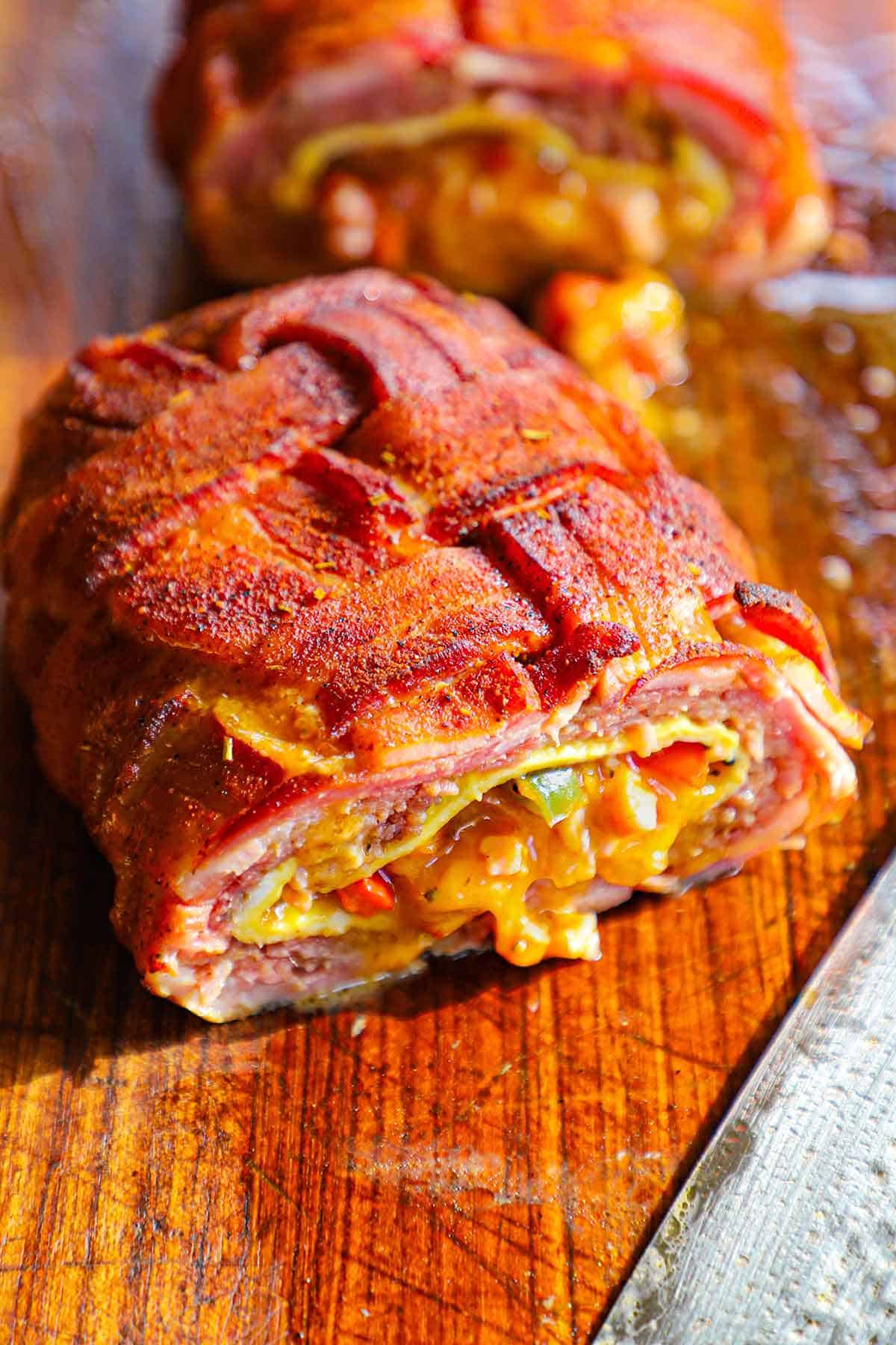 A close-up view of a smoked Western omelette fatty that has been cut in half revealing the melty cheddar cheese, sautéed vegetables, cooked eggs, and juicy sausage, all wrapped in a seasoned bacon weave.