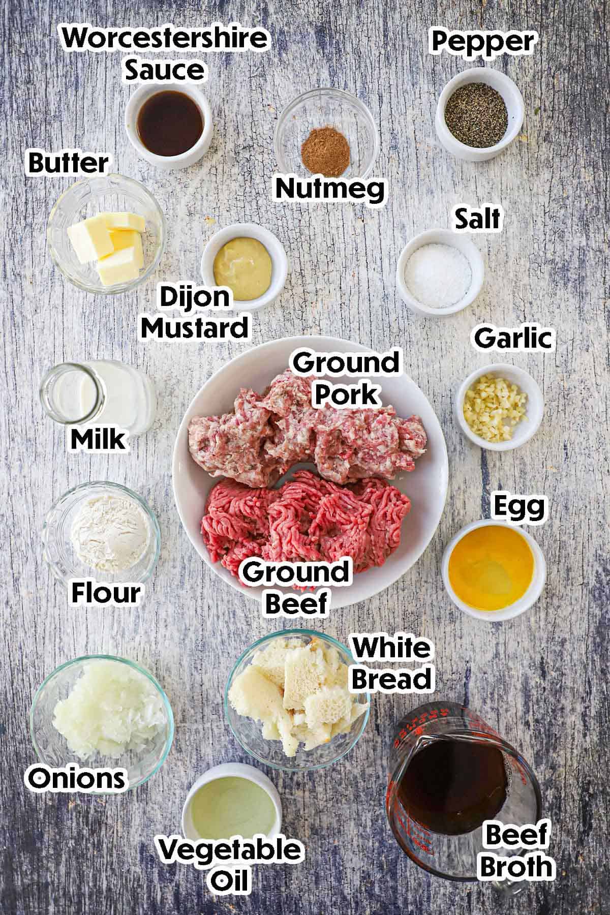 An arrangement of ingredients for Dutch meatballs on a grey wooden background including ground beef, ground pork, white bread, milk, onions, beef broth, garlic, oil, milk, mustard, butter, an egg, and seasonings.