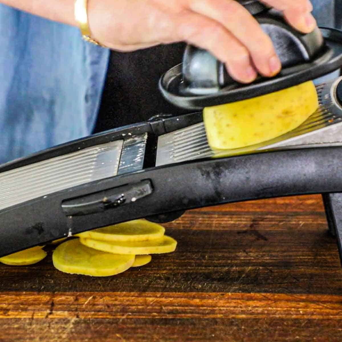 A person using a mandolin to cut gold potatoes into quarter-inch slices on a wooden cutting board.