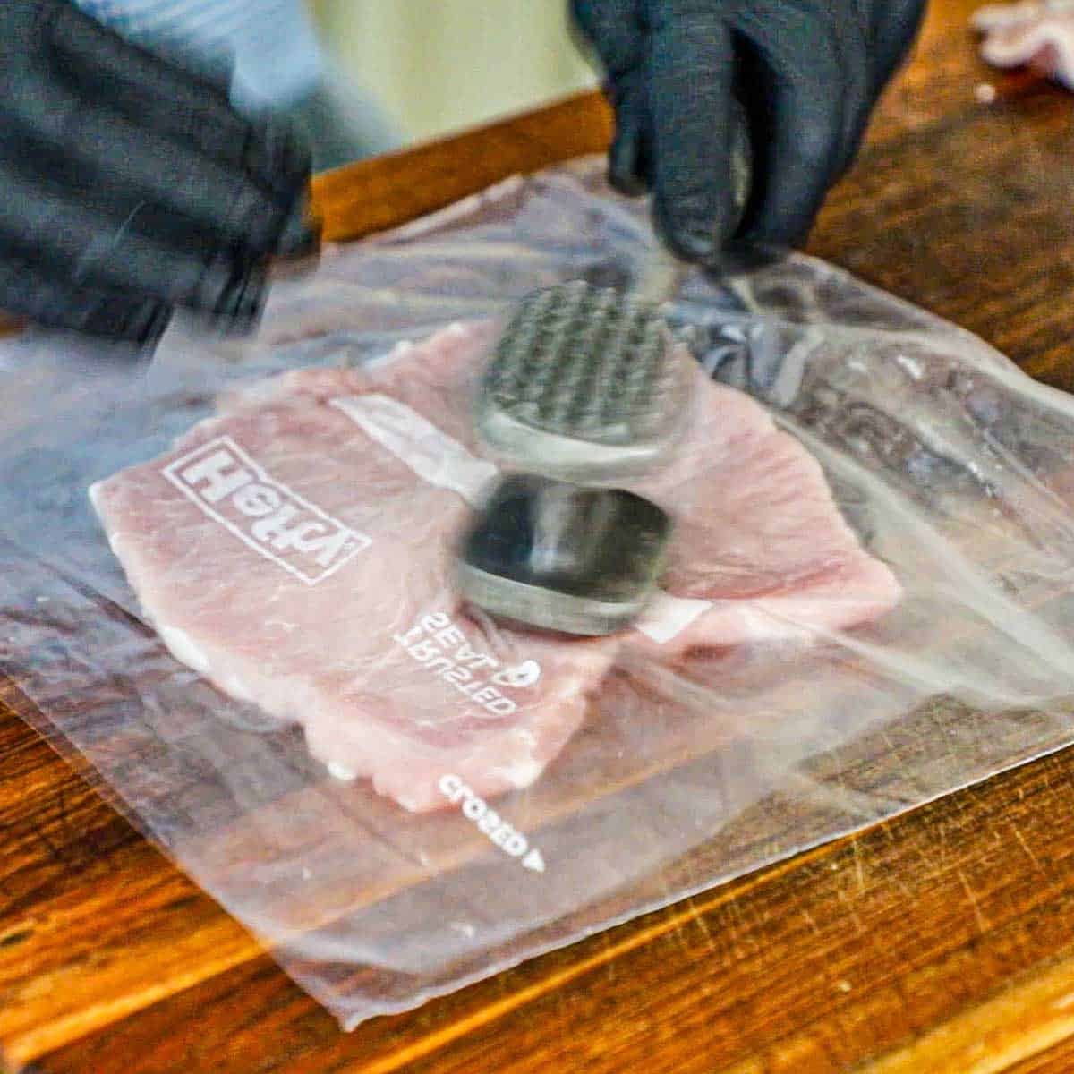 A person using a meat mallet to pound a pork loin chop in between to pieces of plastic wrap on a large wooden cutting board.