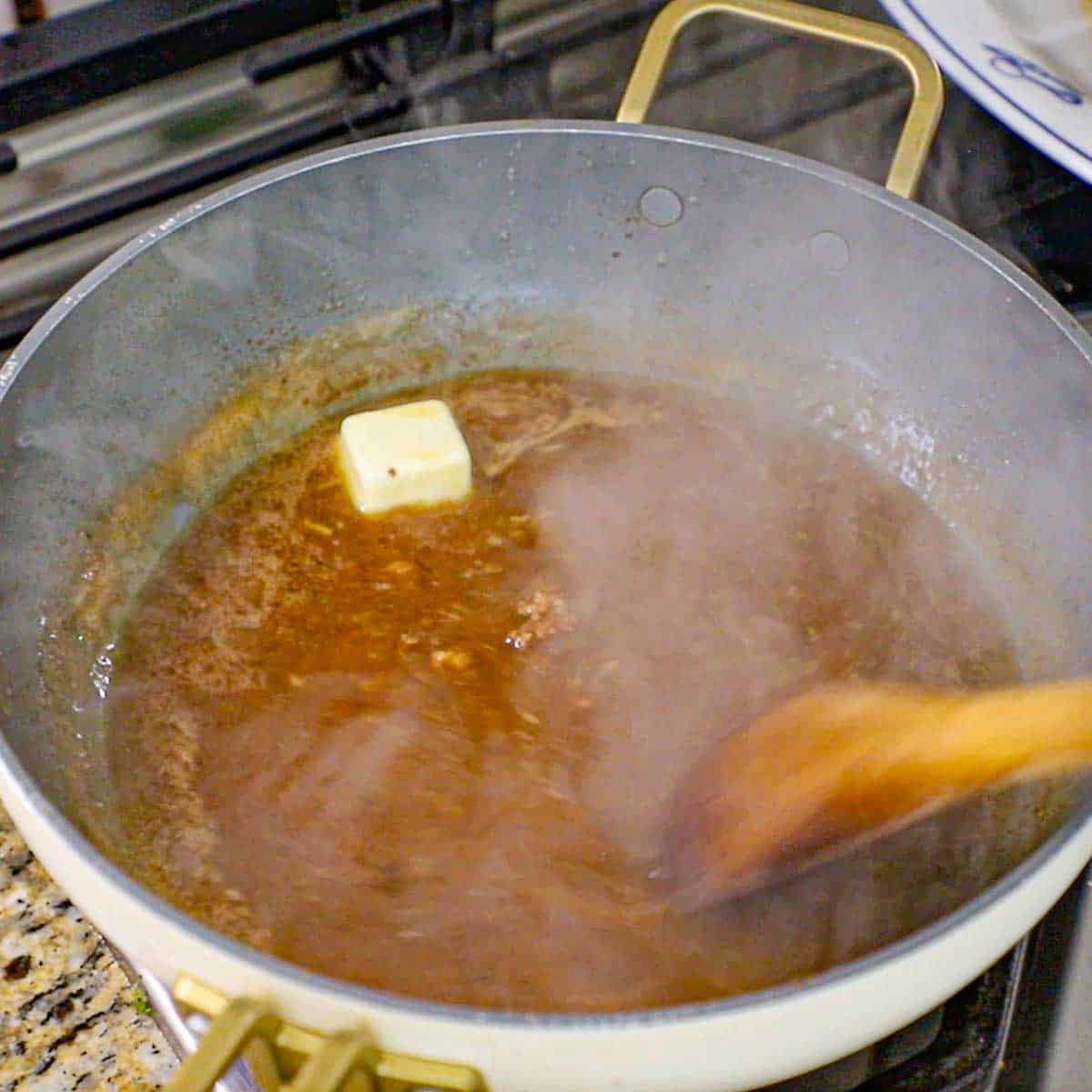 A person using a wooden spoon to stir a beef au jus sauce with a tab of butter that is melting in the sauce in a large skillet on a gas stove.