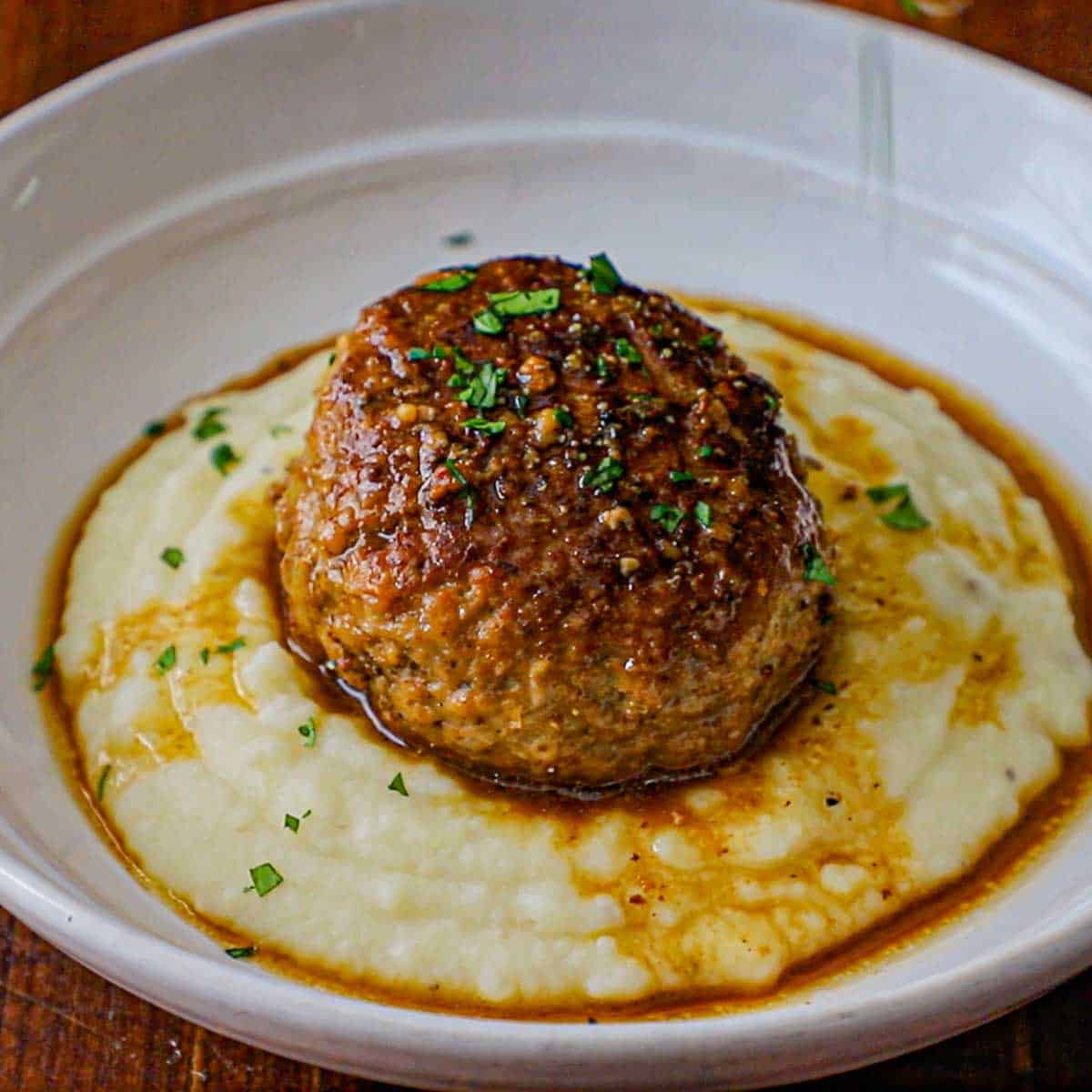 A close-up view of a large cooked Dutch meatball resting on a bed of mashed potatoes that have all been topped with a buttery beef au jus sauce and garnished with chopped Italian parsley.