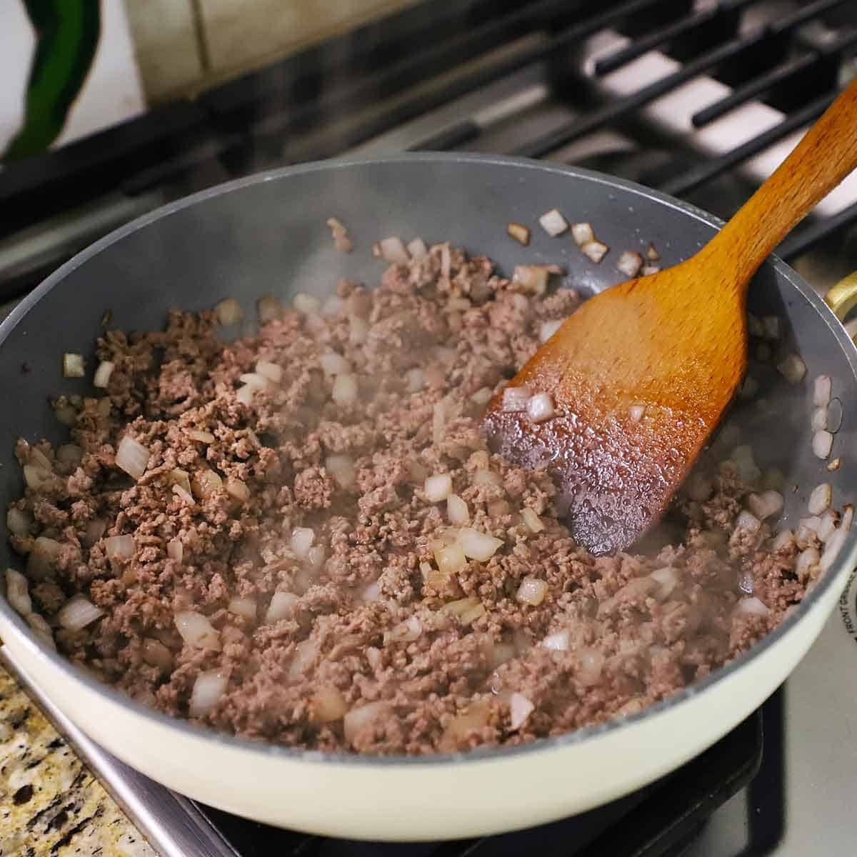 A person using a large wooden spatula to stir cooked ground been with chopped sautéed onions in a large skillet on a gas stove.