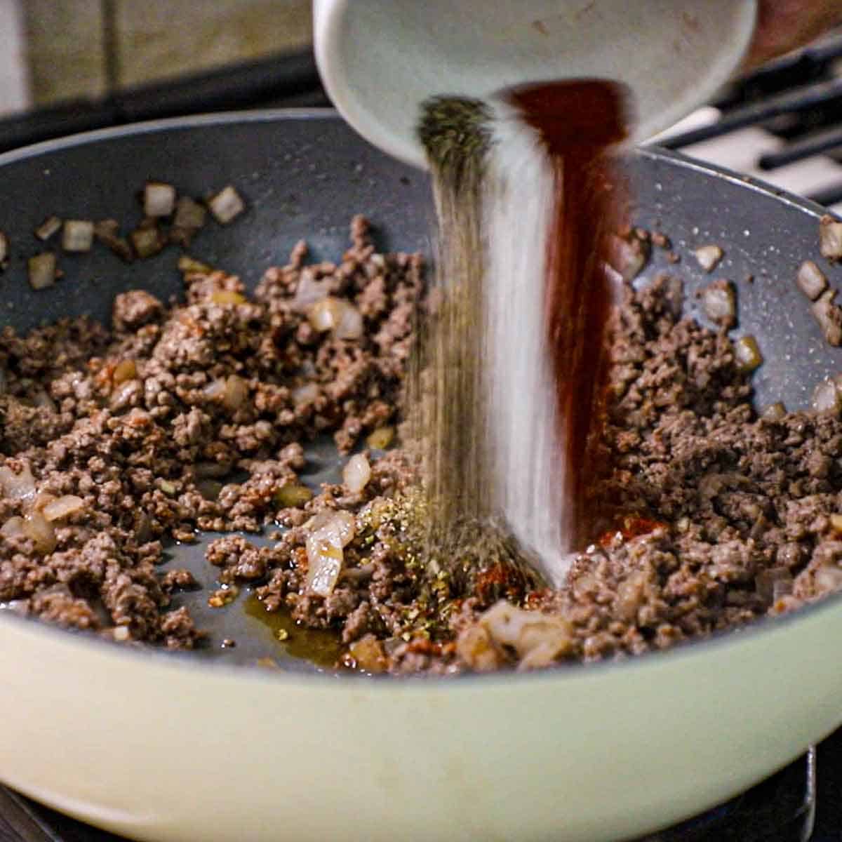 A person dumping seasonings from a small white bowl into a skillet filled with cooked ground beef with tomato paste and sautéed onions.