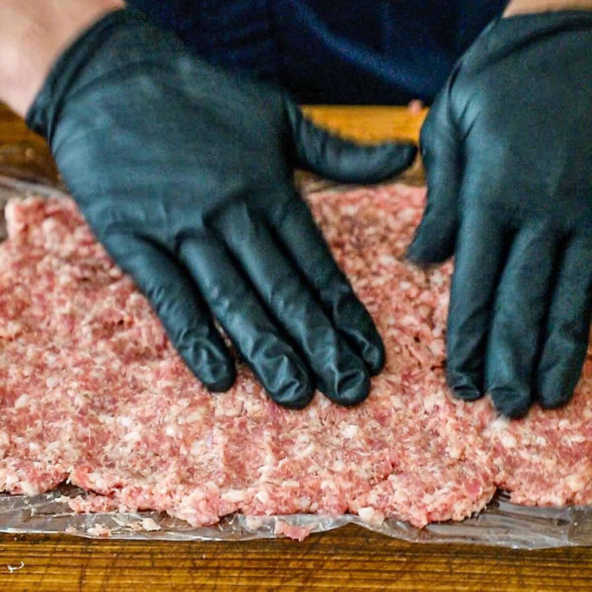 A person wearing black kitchen gloves pressing a thin layer of uncooked breakfast sausage onto a piece of plastic wrap on a large wooden cutting board.