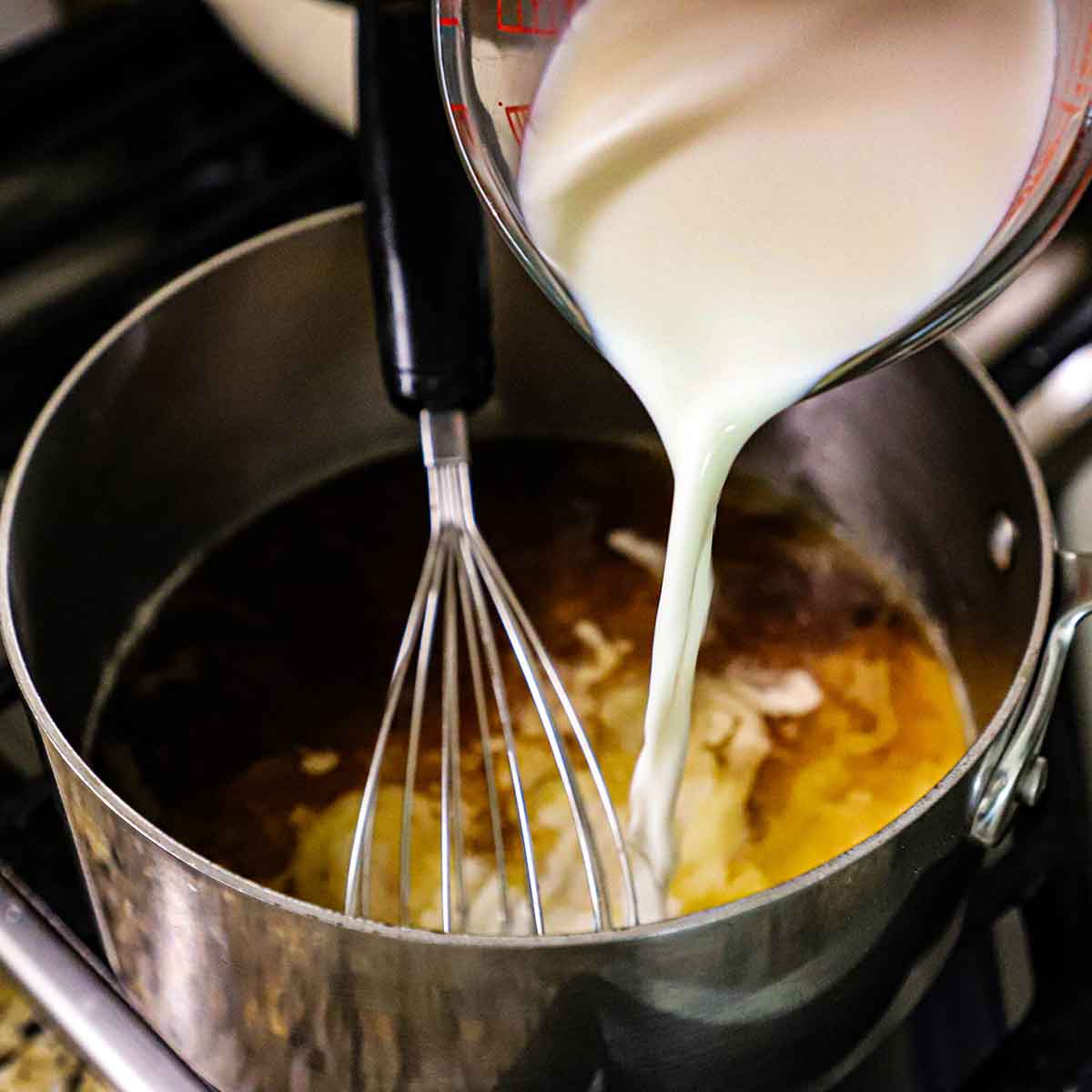 A person pouring whole milk from a glass measuring cup into a steel saucepan with whisk stirring the milk along with beef stock into a roux in the bottom of the pan.