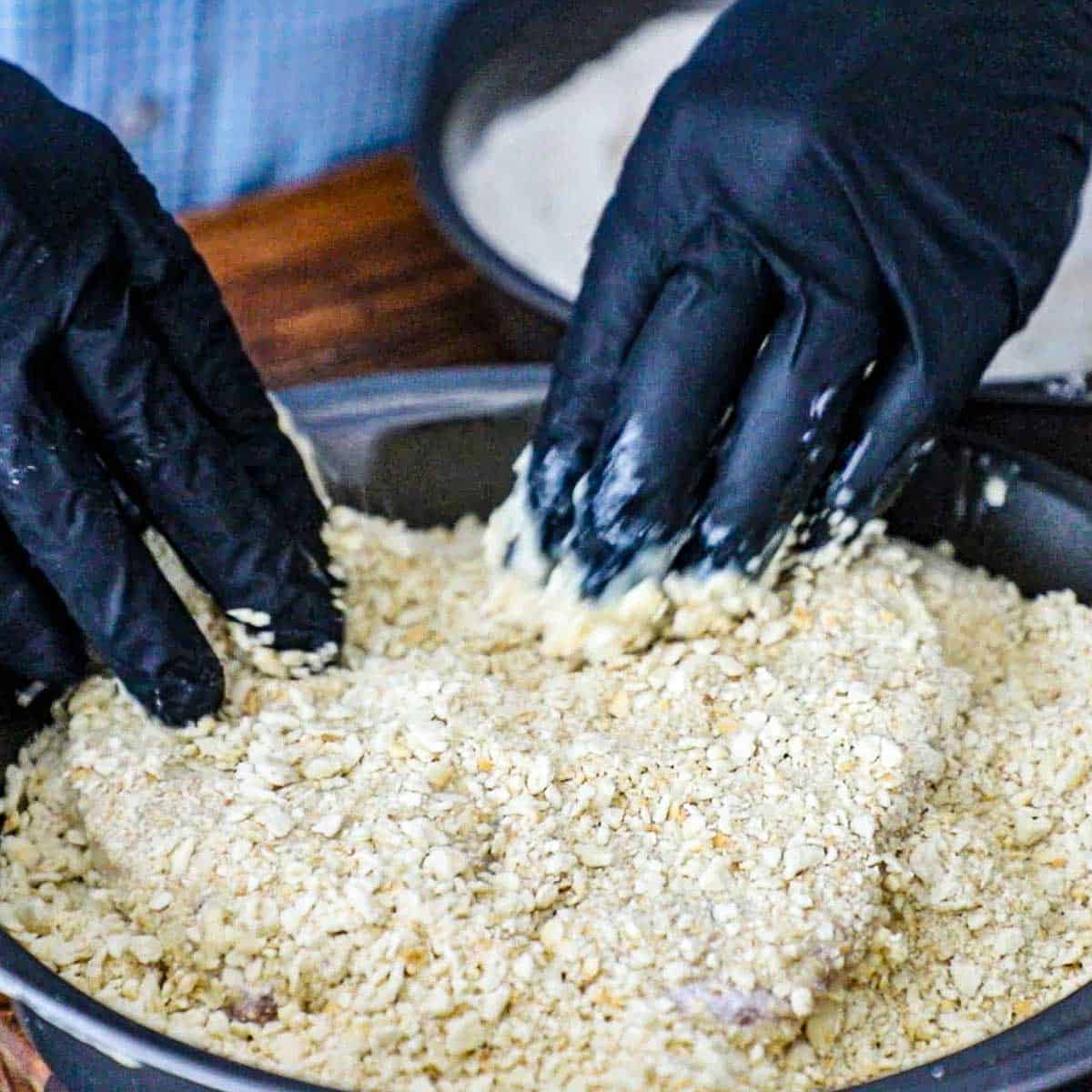 A person wearing black cooking gloves pressing pulverized saltine crackers into a buttermilk-coated pork loin chop in a circular baking pan.