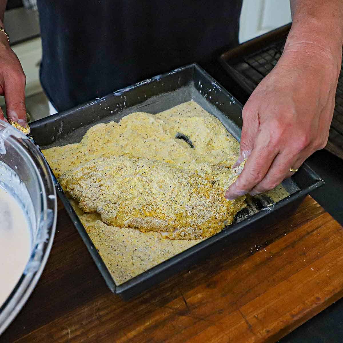 A person using his hands to dredge a fresh catfish filet that was coated in a buttermilk mixture through a seasoned cornmeal and flour mixture in a square baking pan.