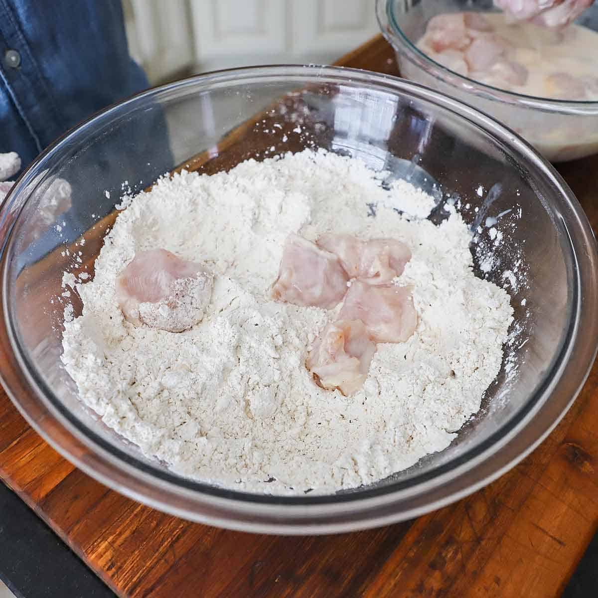 A person placing chunks of chicken breast that have been soaked in a buttermilk and pickle brine mixture into a large glass bowl filled with seasoned flour and cornstarch.