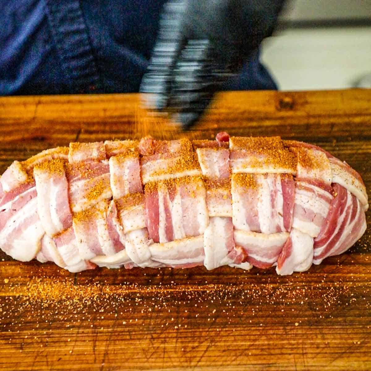 A person sprinkling pork rub over the top of an uncooked Western Omelette Fatty that is encased in a weave of bacon on a large wooden cutting board.