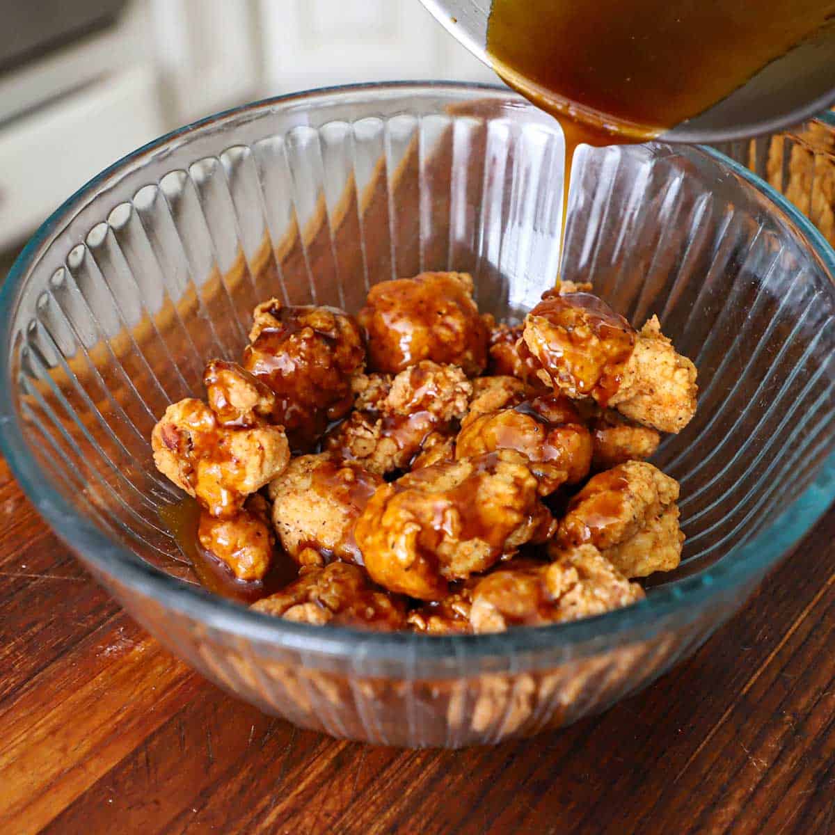 A person pouring a maple Bourbon glaze over crispy boneless chicken wings that are in a glass bowl on a wooden cutting board.