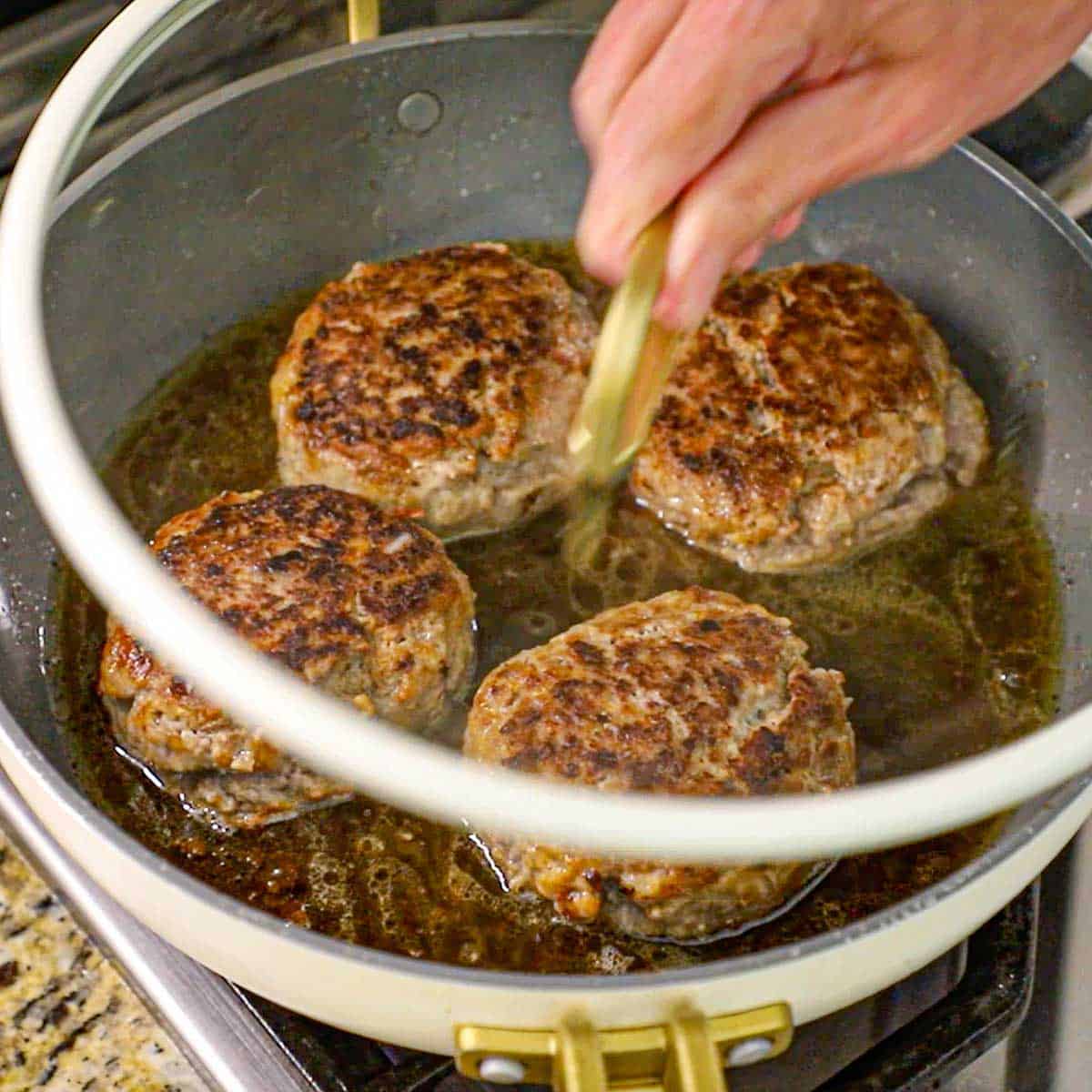 A person placing a glass lid on top of a large non-stick skillet that is holding four large Dutch meatballs in simmering beef stock and melted butter on a gas stove.