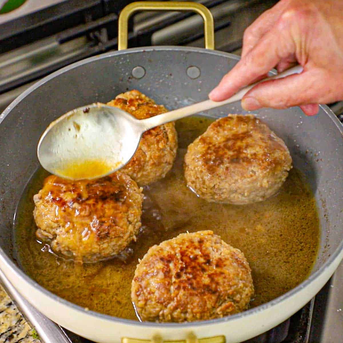 A person using a large spoon to drizzle buttery beef broth from the bottom of a skillet that contains four large Dutch meatballs simmering on a gas stove.